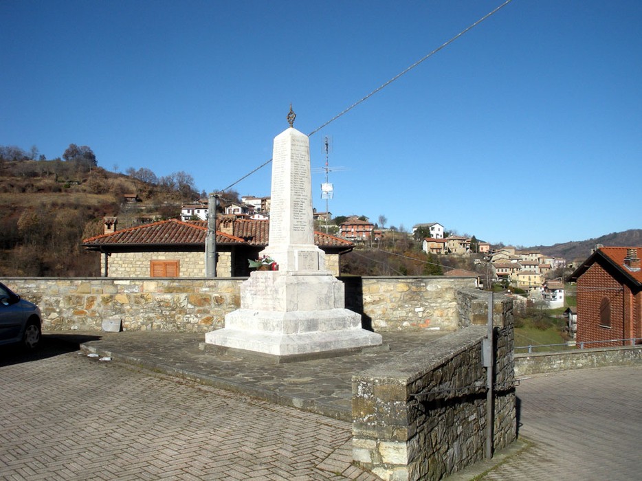Monumento ai caduti di Romagnese, situato in un paesaggio collinare, circondato da abitazioni.
