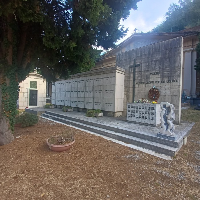 Sacrario con loculi in marmo, croce, iscrizione e corona, in un cimitero alberato.