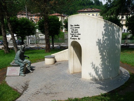 Monumento a Rampinelli con scultura e struttura semicircolare in un'area verde, circondato da alberi e edifici.
