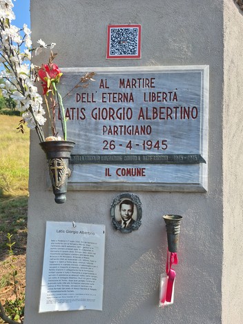 Lapide in marmo su parete esterna, con iscrizione, foto del caduto, fiori e un piccolo lume votivo.