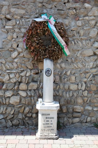 Cippo commemorativo con corona di fiori, situato su un muro di pietra, in omaggio a Anselmo Menabue.