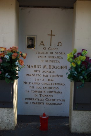 Cippo commemorativo dedicato a Mario M. Ruggeri, con fiori e una croce, in ricordo dei caduti della Seconda Guerra Mondiale.