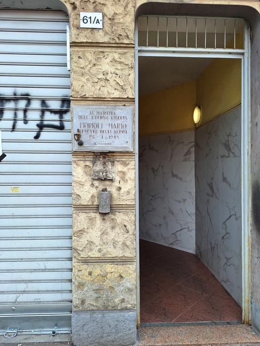 Lapide in marmo con iscrizione, affissa su un muro accanto a un ingresso, vicino a una saracinesca.