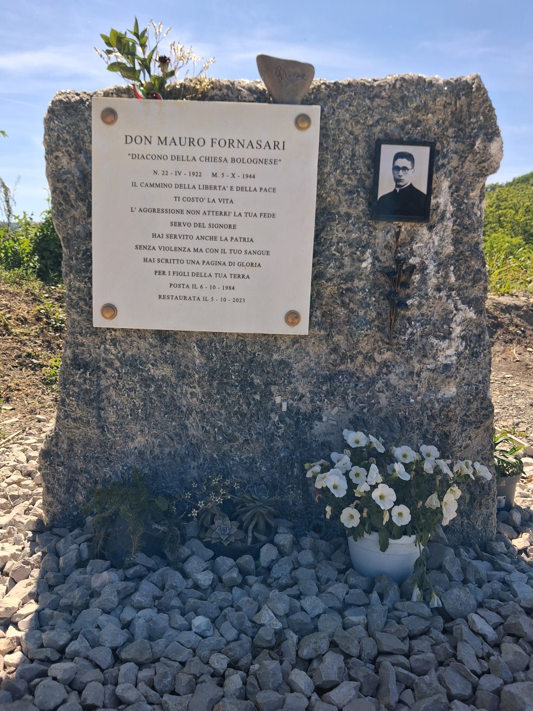 Cippo commemorativo dedicato a Don Mauro Fornasari, con foto e iscrizioni, circondato da fiori e pietre.