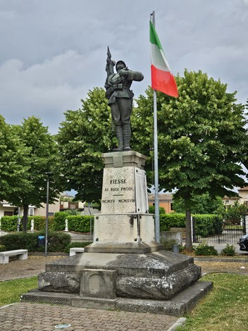 Monumento ai Caduti con statuaria commemorativa e bandiera italiana, circondato da alberi e un'area verde.