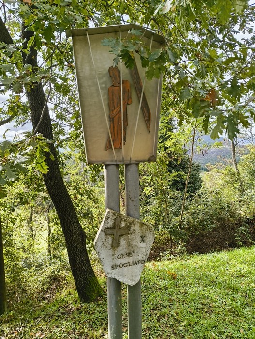 Pannello della Via Crucis di Casaglia, immerso nel verde, con un'immagine di Gesù spogliato.