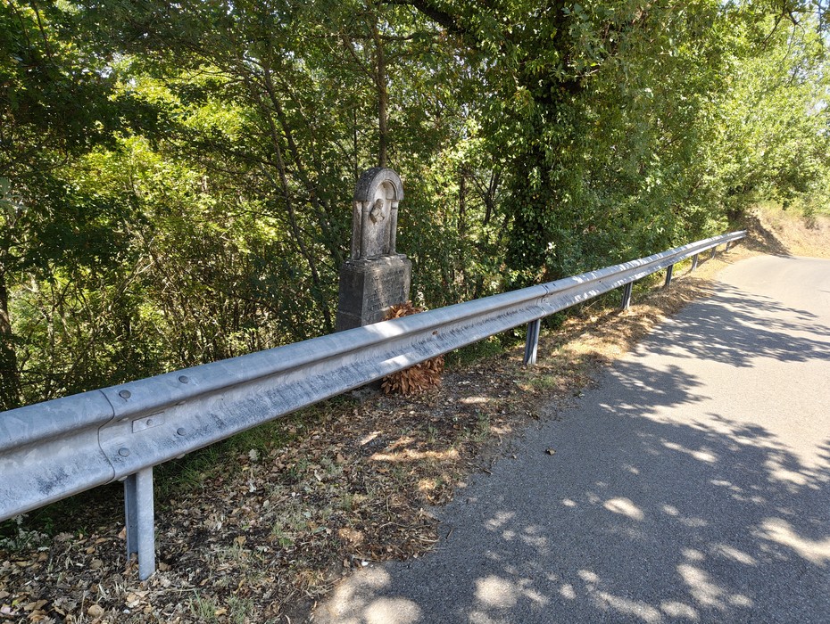 Cippo in pietra con iscrizione, bordo strada tra guardrail e vegetazione.
