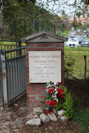 Cippo commemorativo dedicato a Montanari, Gnudi e Benassi, con fiori e pietre in un ambiente verde.