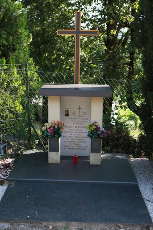 Monumento in memoria di Mario M. Ruggeri, con croce e fiori, immerso nel verde.
