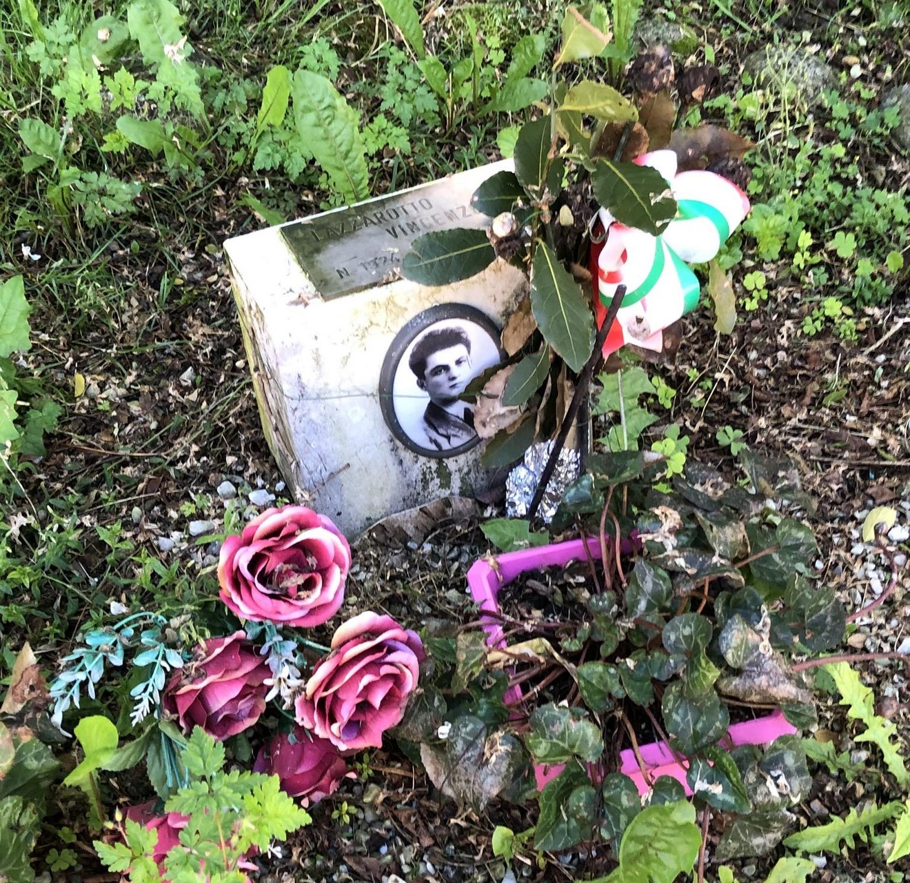 Cippo commemorativo con foto e fiori, a ricordo di Lazzarotto, immerso nella vegetazione.
