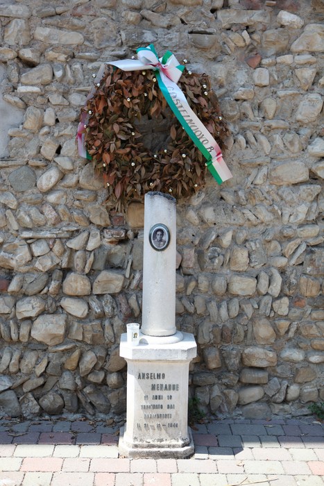 Cippo commemorativo con corona di fiori, situato su un muro di pietra, in omaggio a Anselmo Menabue.