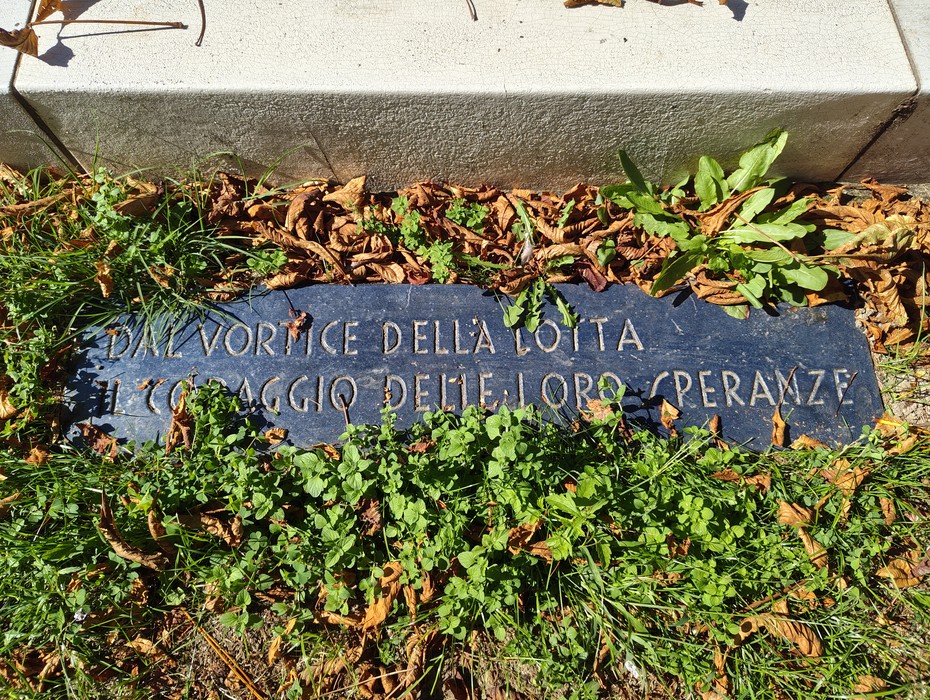 Lastra in pietra scura con iscrizione, posata a terra tra erba e foglie secche, ai piedi di un gradino.