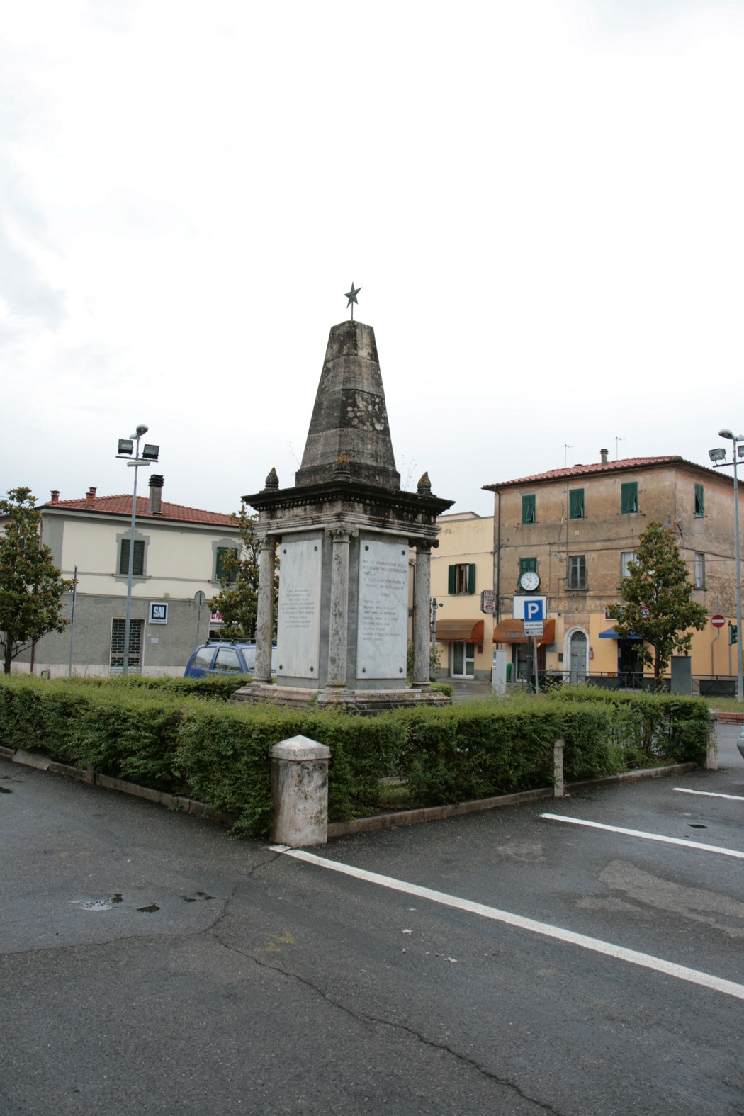 Monumento ai caduti di Perignano, un obelisco commemorativo situato in una piazza circondata da verde.