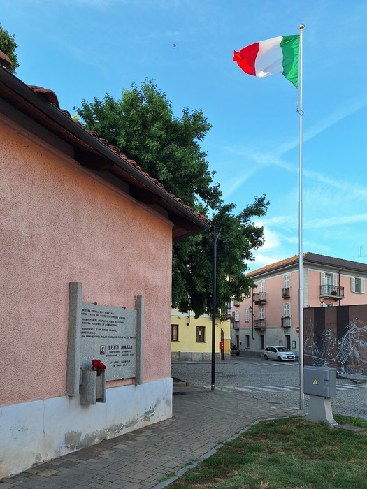 Lapide commemorativa all'aperto con il tricolore italiano e un edificio colorato sullo sfondo.