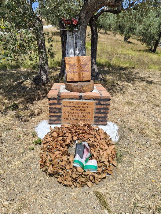 Cippo in mattoni con lapide e libro in pietra scolpito, sormontato da un ulivo, corona di foglie secche alla base.