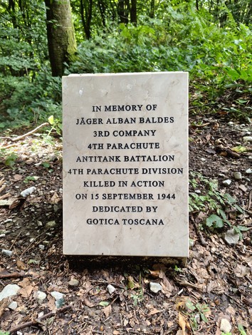 Cippo commemorativo dedicato a Jäger Alban Baldes, situato in un'area boschiva, ricco di vegetazione.