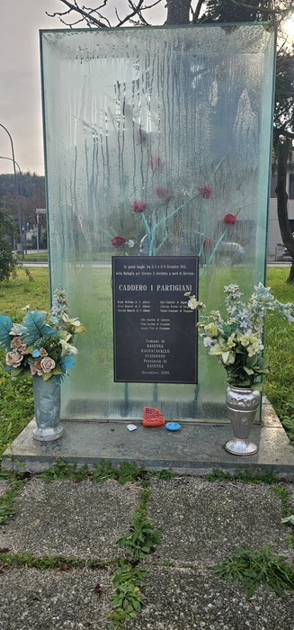 Monumento commemorativo trasparente con fiori e lastre in vetro, dedicato a Beltrami e altri.