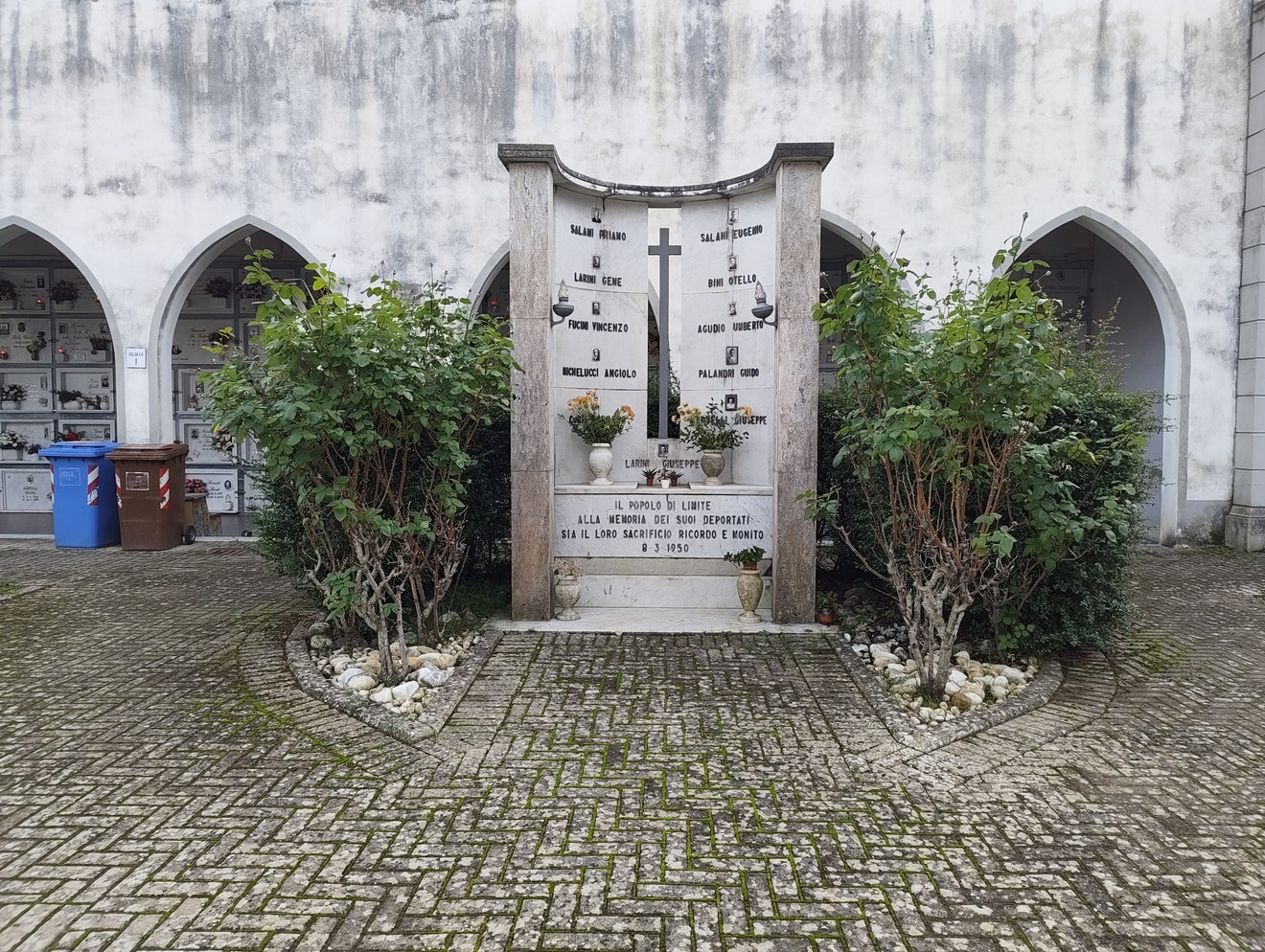 Sacrario dei deportati con monumento commemorativo circondato da vegetazione e muretti in un contesto sobrio e rispettoso.