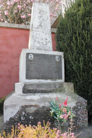Cippo commemorativo con iscrizioni e decorazioni floreali, circondato da vegetazione e fiori.
