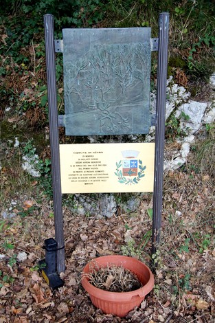 Targa commemorativa su supporto metallico, circondata da vegetazione e un vaso di fiori.