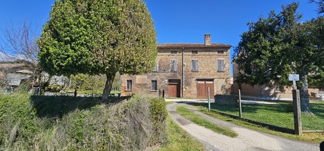 Lapide commemorativa immersa in un paesaggio naturale con una casa storica sullo sfondo.