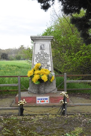 Monumento dedicato a Benassi, con fiori e scritta commemorativa, immerso nel verde.