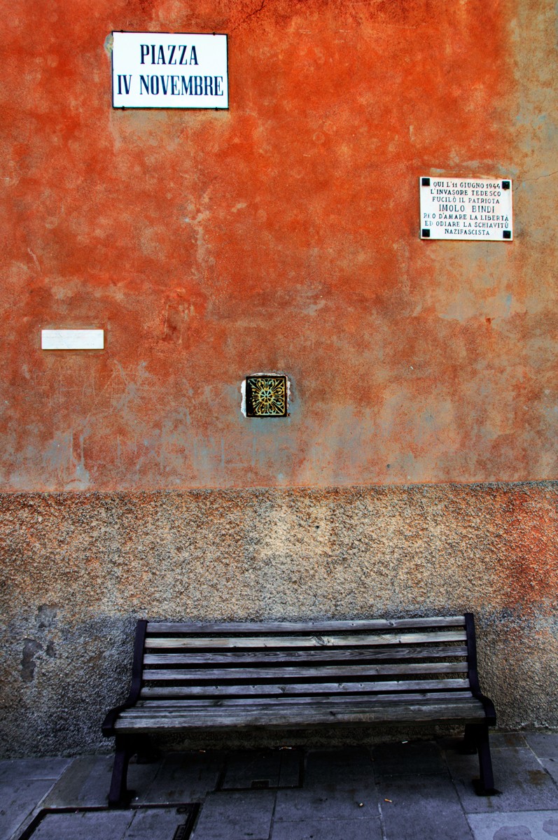 Lapide commemorativa su un muro arancione con panchina in piazza IV Novembre.