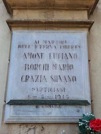 Lapide in marmo con iscrizione, fissata su parete in pietra, con fiori deposti sotto.