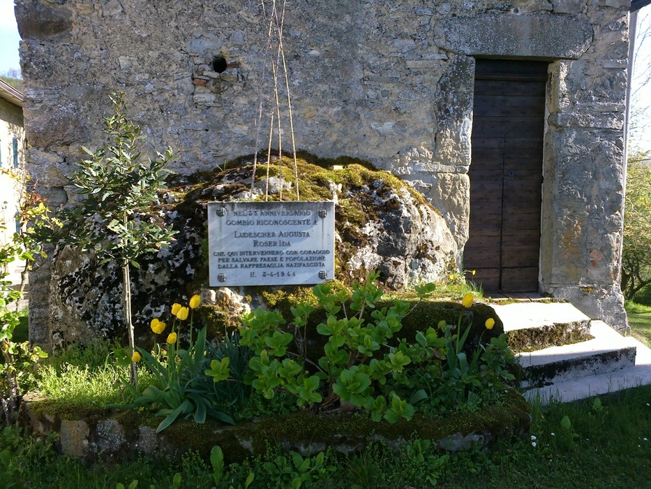 Lapide commemorativa a Ludescher e Roser, situata presso un edificio storico con vegetazione circostante.