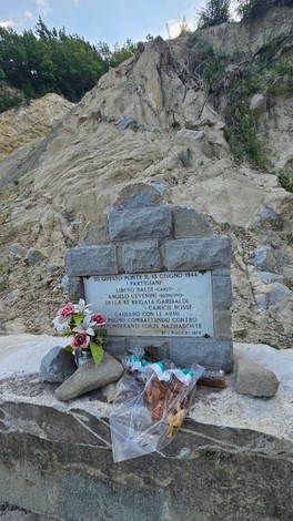 Monumento commemorativo a Baldi e Cevenini, situato in un ambiente naturale montano, con fiori e decorazioni.