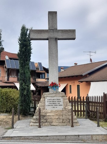 Monumento "Croce ai Martiri di Cumiana" con un'alta croce e decorazioni floreali, circondato da edifici e piante.