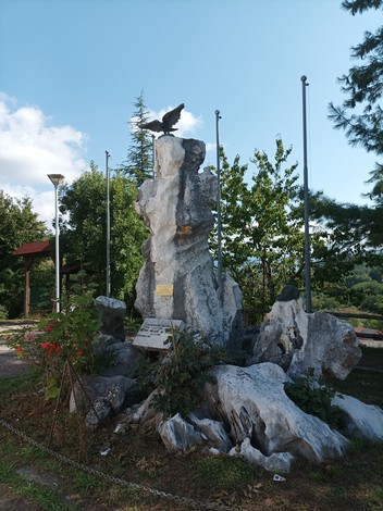 Monumento ai caduti in guerra con una scultura di un'aquila su una base di roccia, circondato da alberi e bandiere.