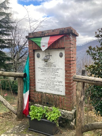 Monumento a Bruno Stefani, con bandiera italiana, situato in un'area verde tra gli alberi.