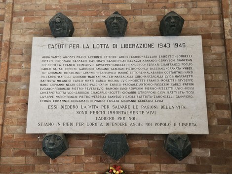 Lapide in marmo su parete, con iscrizione e tre volti scolpiti in rilievo nella parte superiore.