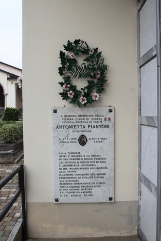 Lapide commemorativa in memoria di Antonietta Piantoni, adornata da una corona di fiori.