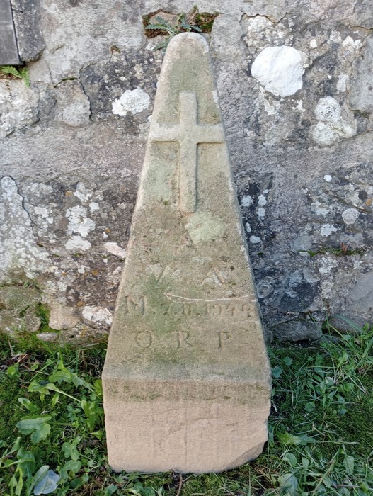 Cippo commemorativo di Videschi, con superficie ruvida e decorato da una croce, immerso nella vegetazione.
