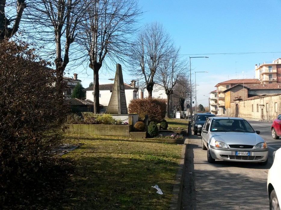 Monumento ad Arzani circondato da alberi e vegetazione, con veicoli in transito sulla strada vicina.