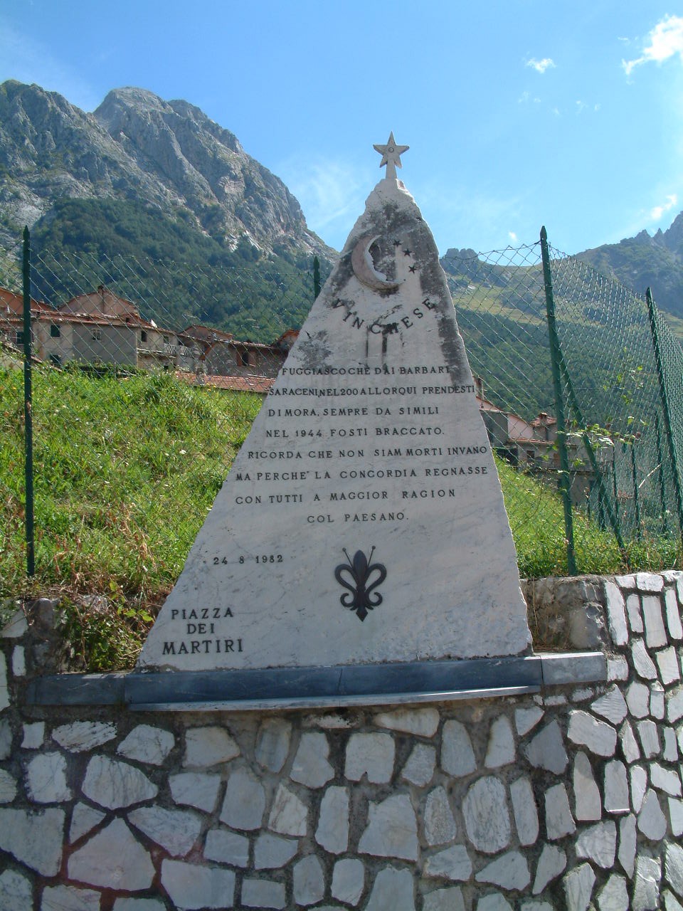 Cippo commemorativo dedicato ai martiri, situato in Piazza dei Martiri, circondato da un paesaggio montano.