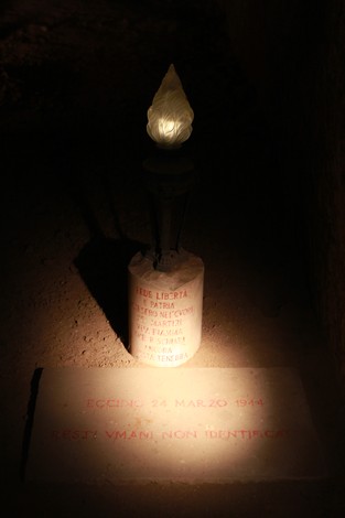 Memoriale con lumino, incisi nomi e data in ombra, simbolo di commemorazione e riflessione storica.