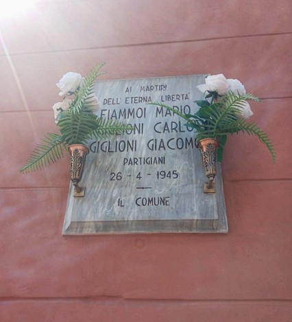 Lapide in marmo con iscrizione, affissa a una parete, ornata da due vasi con fiori bianchi e felci.