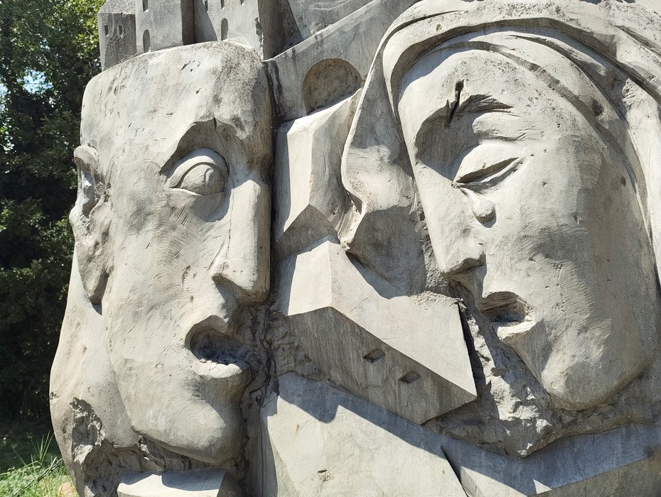 Dettaglio scultoreo in pietra del Monumento alla Libertà, con due volti umani scolpiti ad alto rilievo.