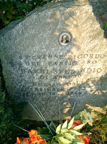 Cippo commemorativo con foto e incisioni, circondato da fiori, in un'area tranquilla.