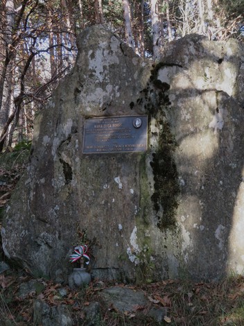 Targa commemorativa su una roccia nel bosco, circondata da alberi e decorata con un nastro tricolore.