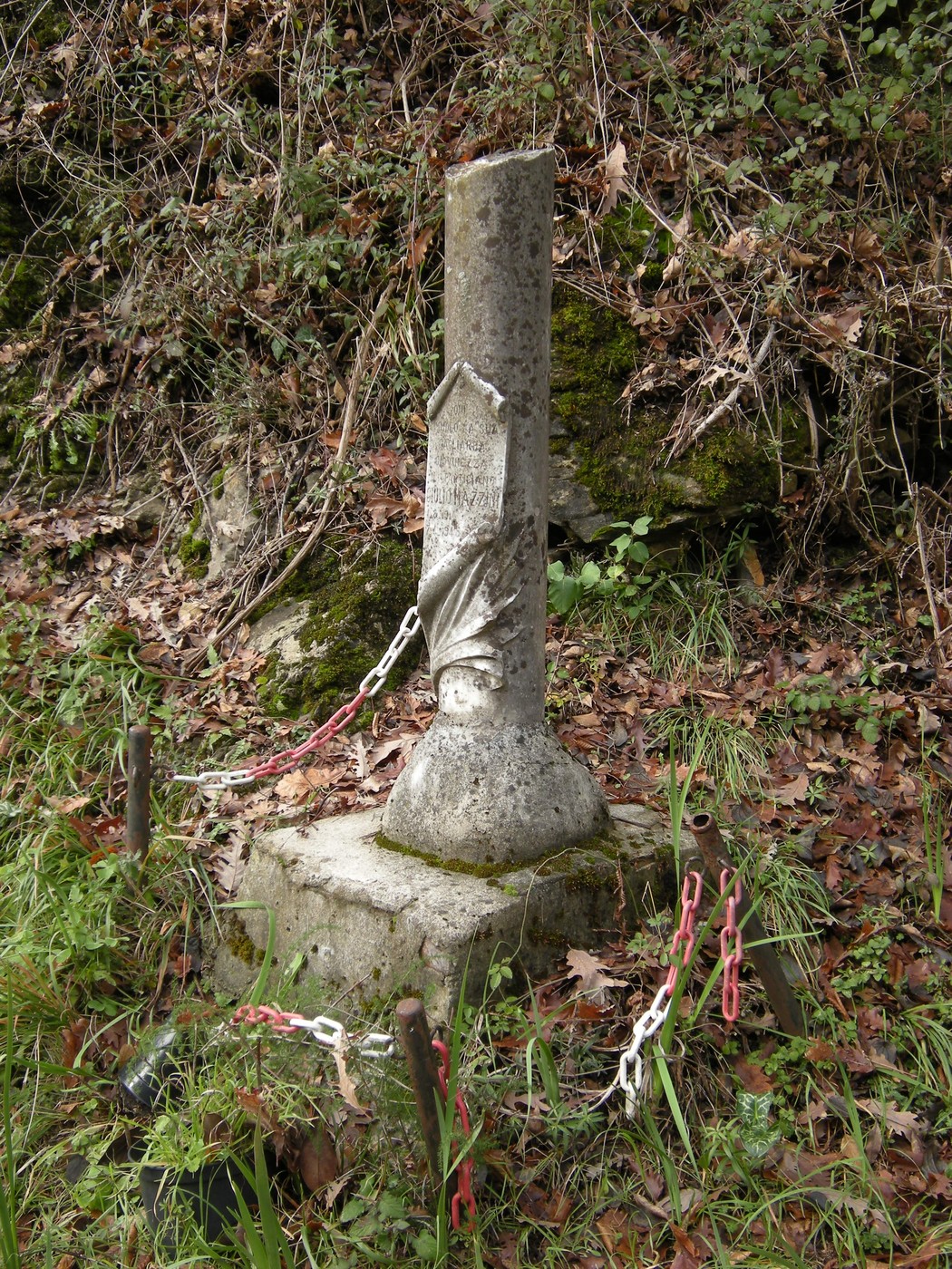 Cippo commemorativo dedicato a Giuseppe Mazzini, circondato da vegetazione e catene, in un contesto naturale.