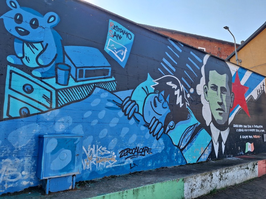 Murale su parete esterna con un orso di peluche, una figura dormiente e un ritratto maschile con iscrizione.