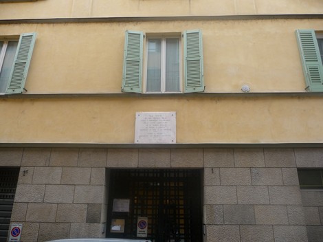 Lapide commemorativa affissa su una parete gialla, sotto finestre con persiane verdi.
