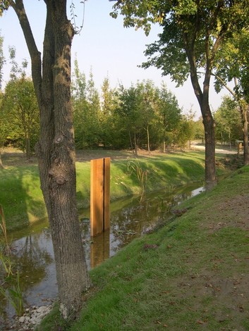 Una stele di metallo emerge tra gli alberi in un paesaggio verdeggiante vicino a un canale d'acqua.