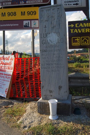 Cippo commemorativo dedicato a Gianello, con scritte e segnaletica stradale sullo sfondo.