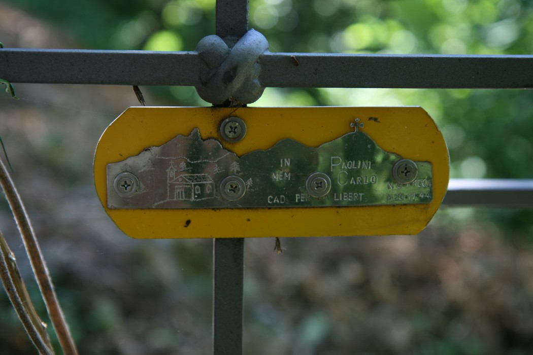 Croce commemorativa con targhetta in metallo incisa, circondata da vegetazione.