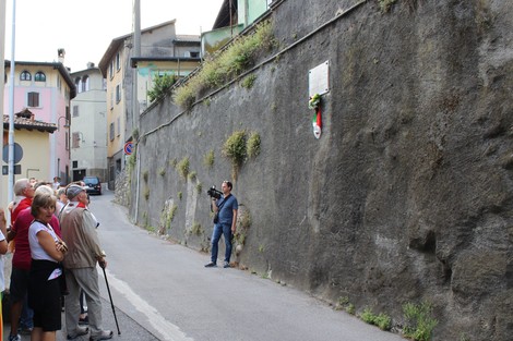 Cerimonia in strada con una lapide commemorativa su un muro roccioso, persone assistere all'evento.